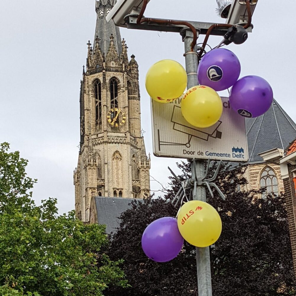 Pilaar met 2 camera's van de Gemeente Delft die is versierd met ballonnen van Piratenpartij Delft en STIP, met op de achtergrond de toren van de nieuwe kerk.